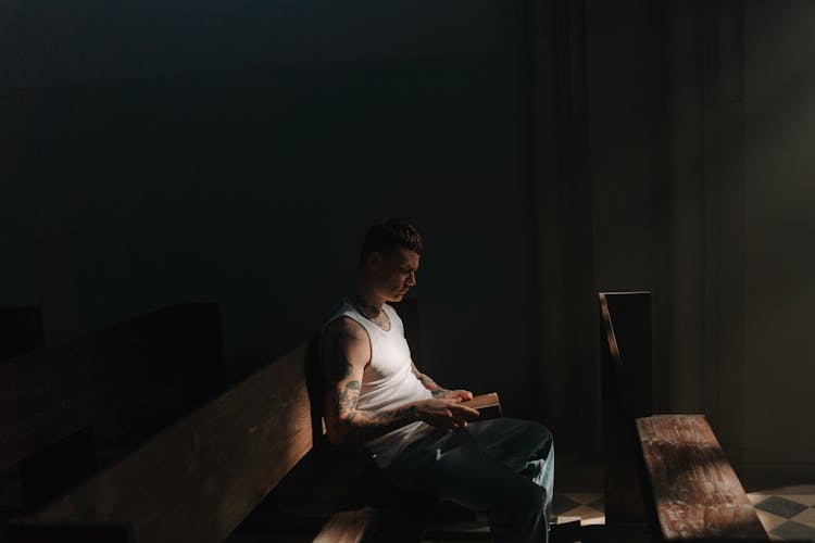 A Side View Of A Man In White Tank Top Sitting On A Pew While Reading A Book