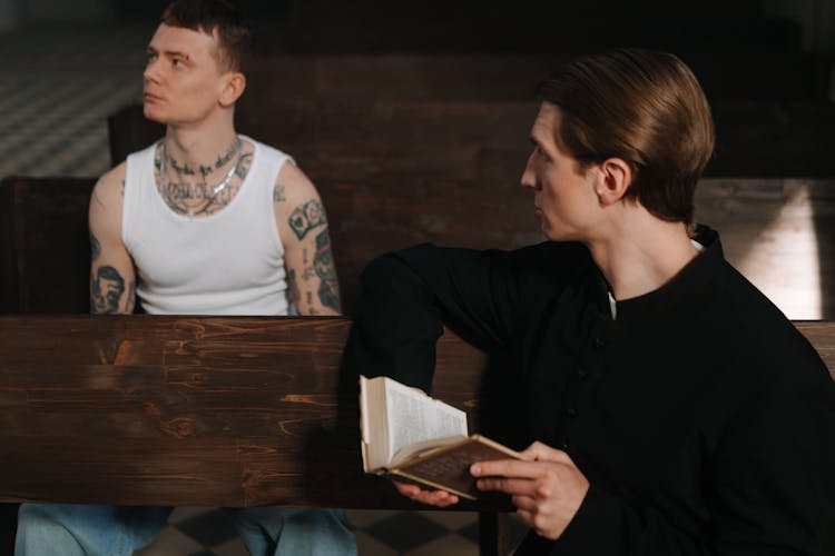 A Priest Holding A Bible While Looking At The Tattooed Man In White Tank Top