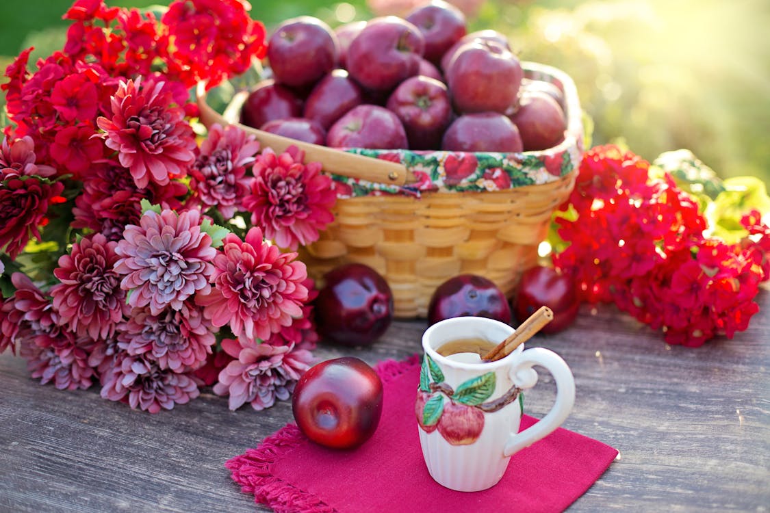 A Basket of Red Apples on a Wooden Table with Flowers Beside a Mug with Drink