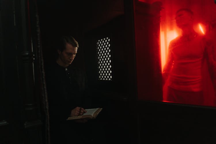 Priest Sitting Inside A Confession Box  With A Bible