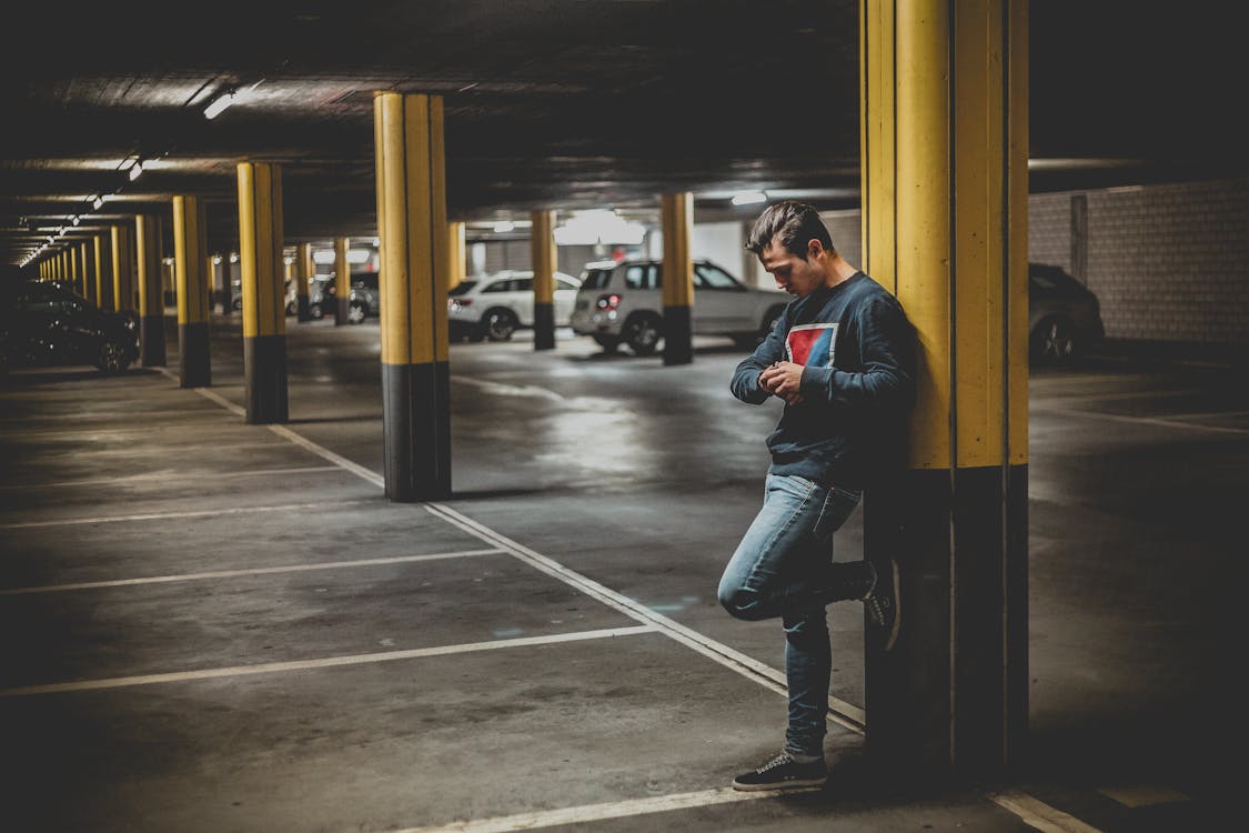 Man in Dark-gray Crew-neck Sweater Standing Leaning to Post in Parking Lot