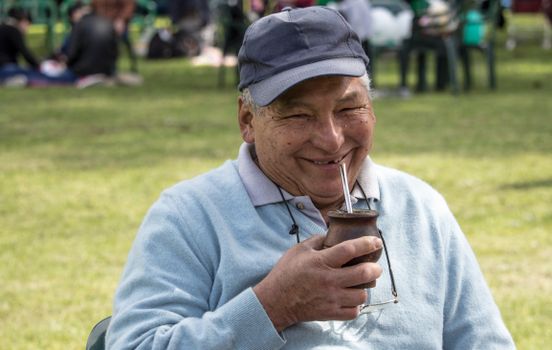 A joyful elderly man sipping yerba mate outdoors, wearing glasses and a cap, in a park setting.