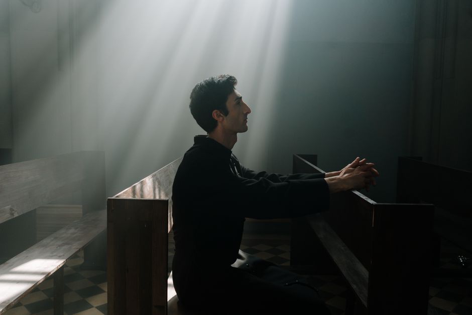 A man sits praying in sunlight inside a dramatic church setting.