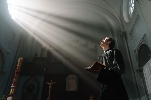 A priest prays holding a Bible under radiant sunrays in a cathedral interior.