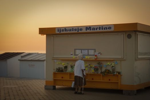 An elderly woman stands by a floral display at a kiosk during sunset.