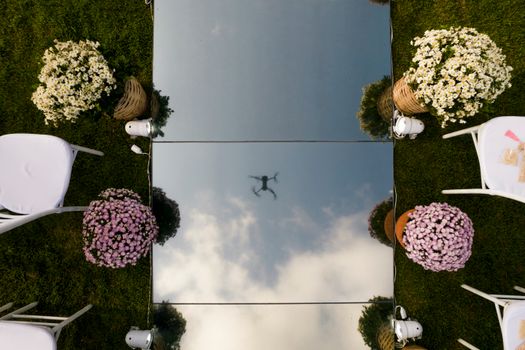 A drone's reflection seen on mirror surface surrounded by flowers and chairs outdoors.