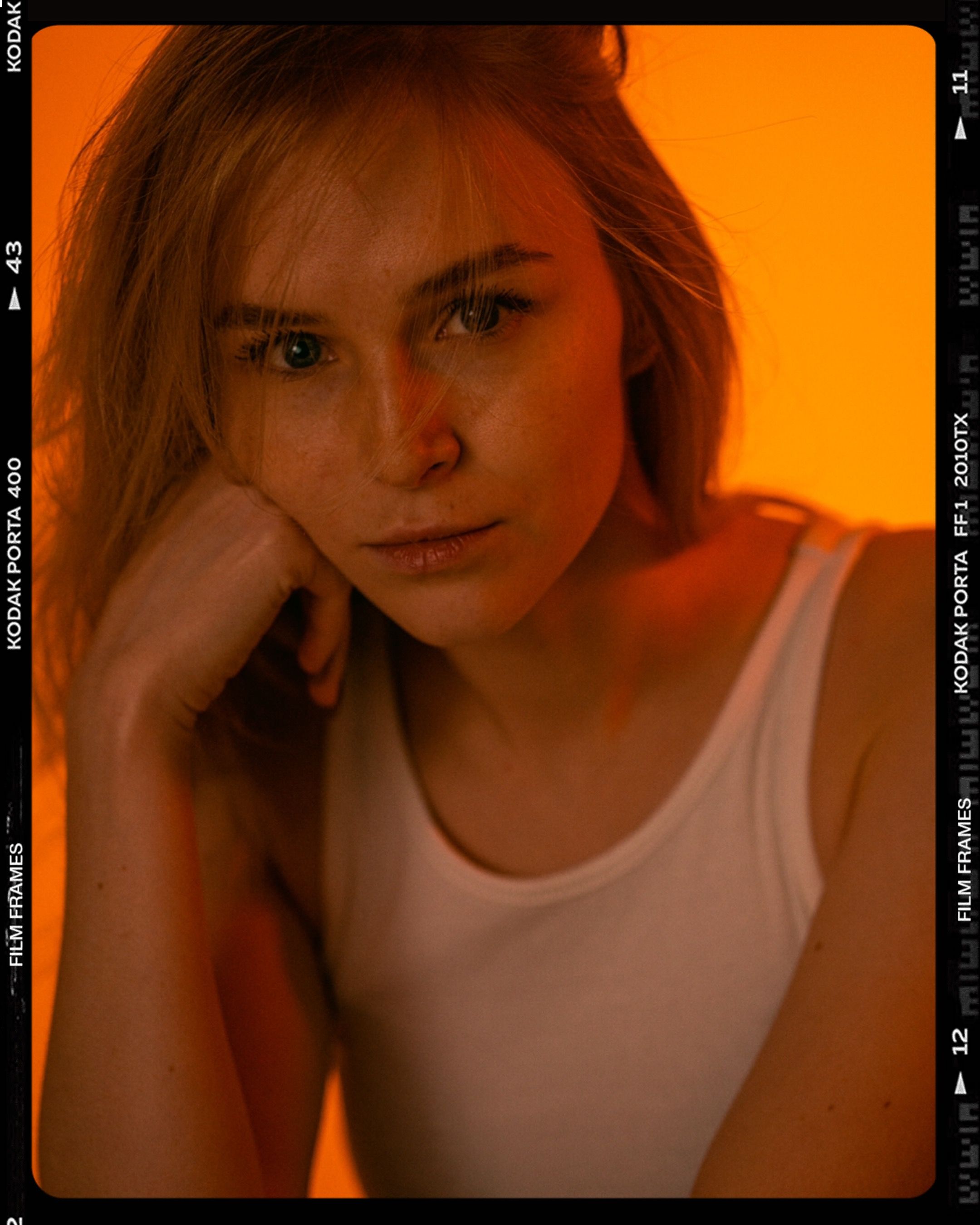 A striking portrait of a woman in a white tank top, featuring a warm orange background.