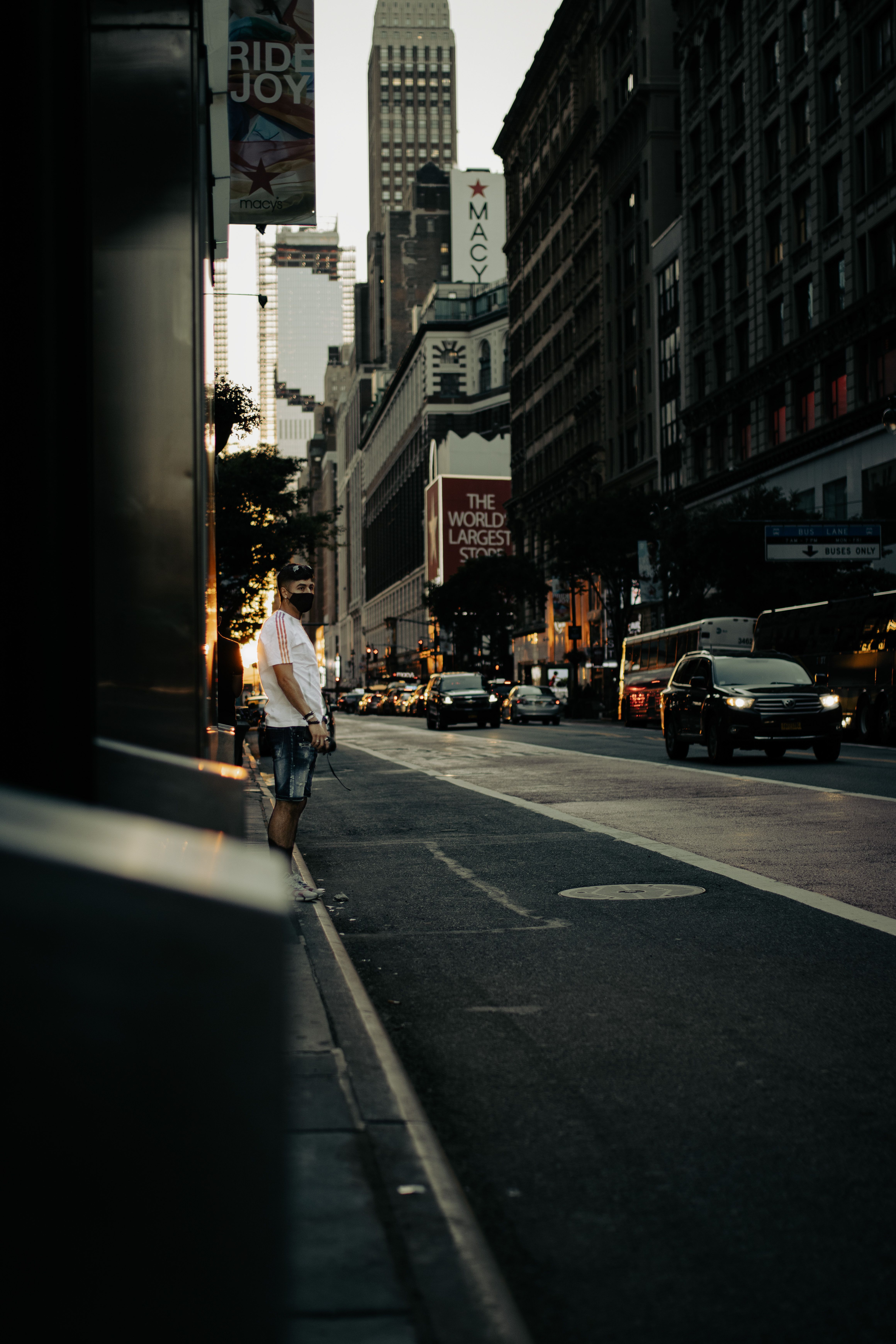 A man in shorts stands on a New York City street at dusk, surrounded by cars and towering buildings.