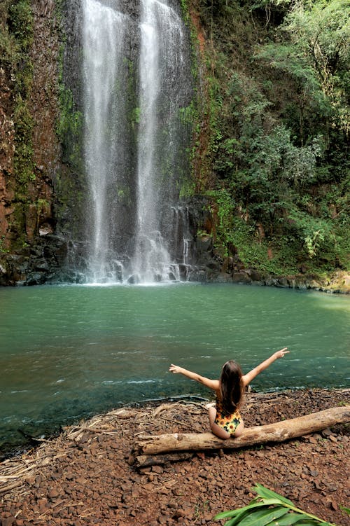 Free A Woman in Swimsuit Sitting on a Tree Log while Facing the Waterfalls Stock Photo