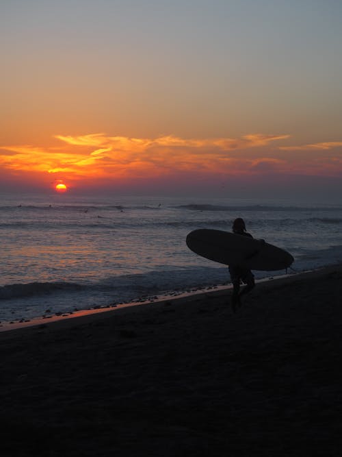 Free Silhouette of a Person Holding a Surfboard on the Beach during Sunset Stock Photo