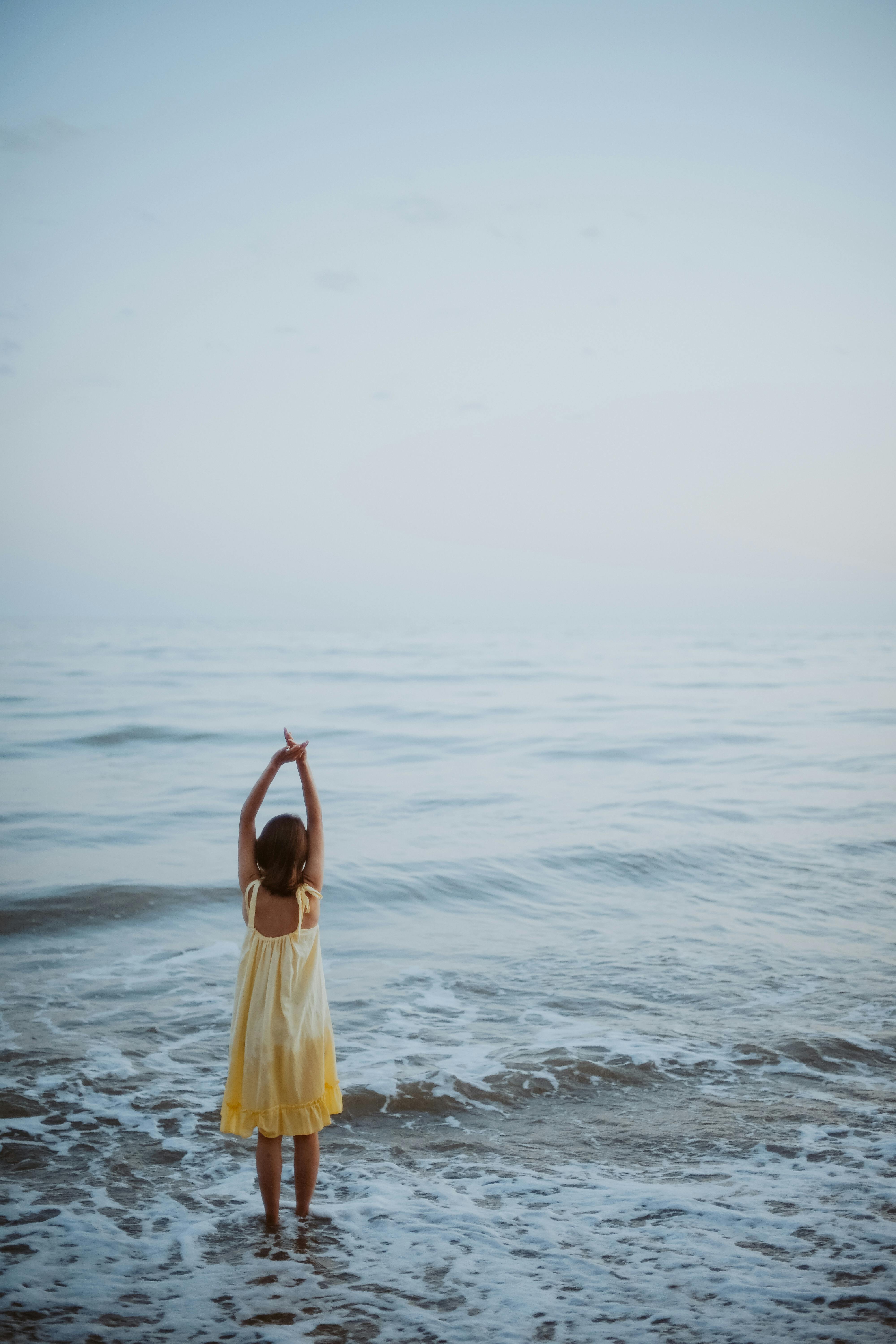A woman in a yellow dress stands at the ocean's edge with arms raised, embracing the serene seascape.