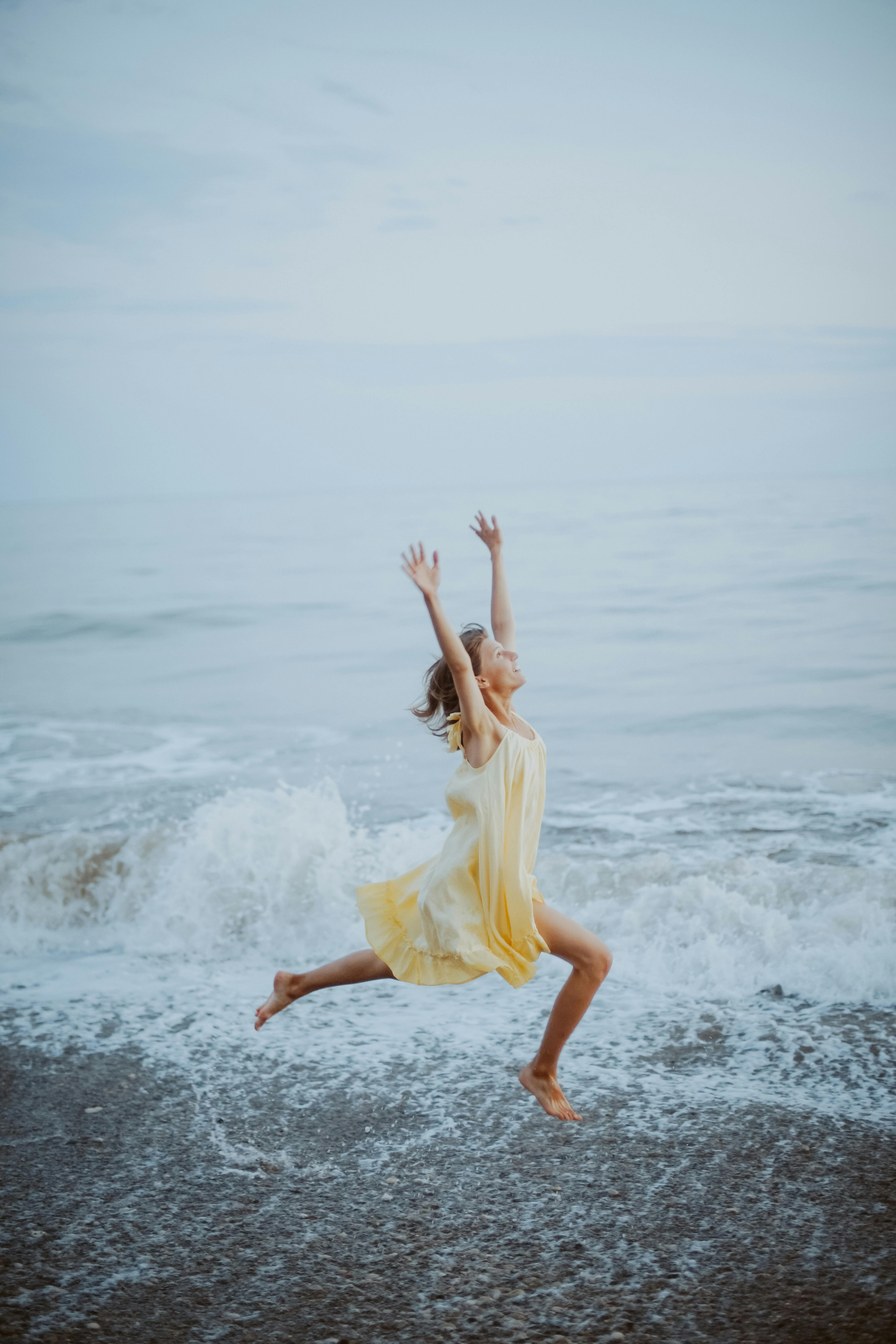 A woman in a yellow dress joyfully jumps on a serene beach with gentle waves.