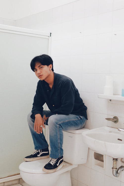 Man in Black Dress Shirt and Blue Denim Jeans Sitting on White Ceramic Toilet Bowl