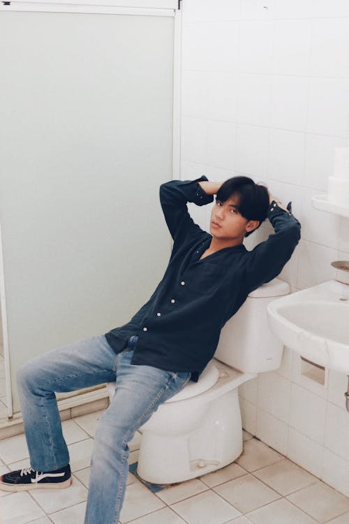 Man in Blue Dress Shirt and Blue Denim Jeans Sitting on White Ceramic Toilet Bowl