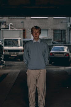 A young man stands in an urban alley, wearing a sweater and chinos. Moody atmosphere.