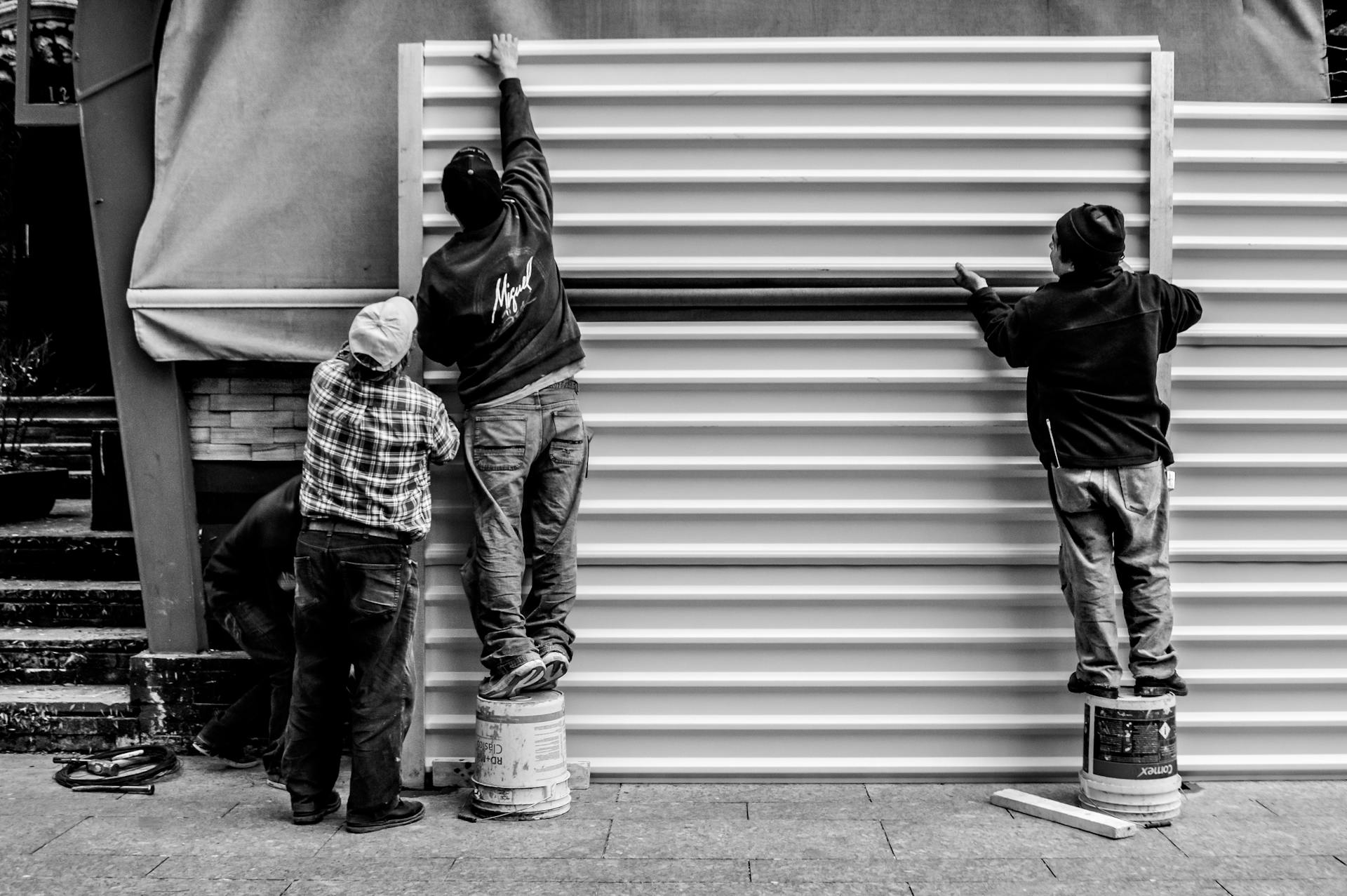 Black and white photo of three workers assembling metal panels outdoors, standing on buckets.