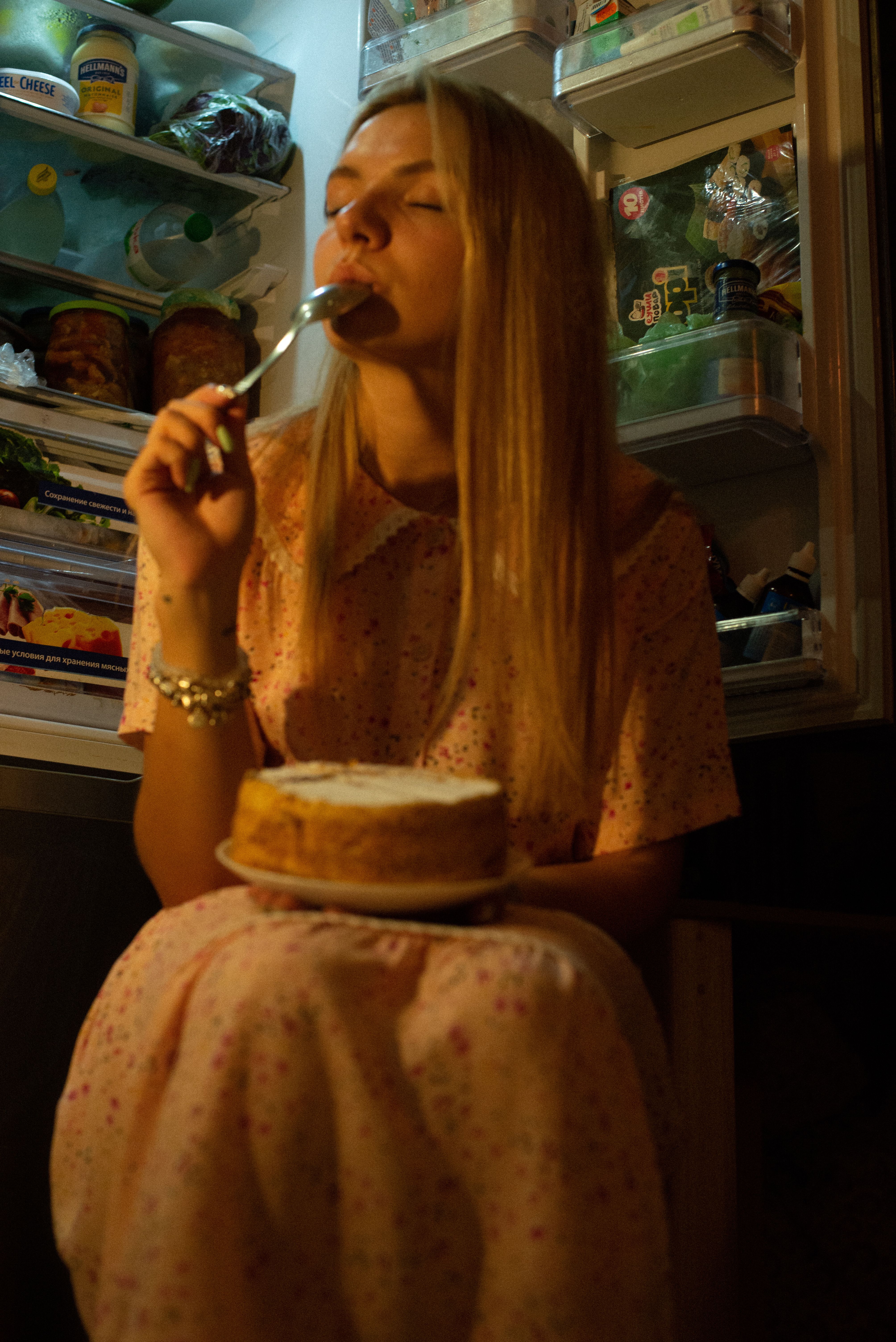 Someone eating a slice of cake in front of an open fridge at night