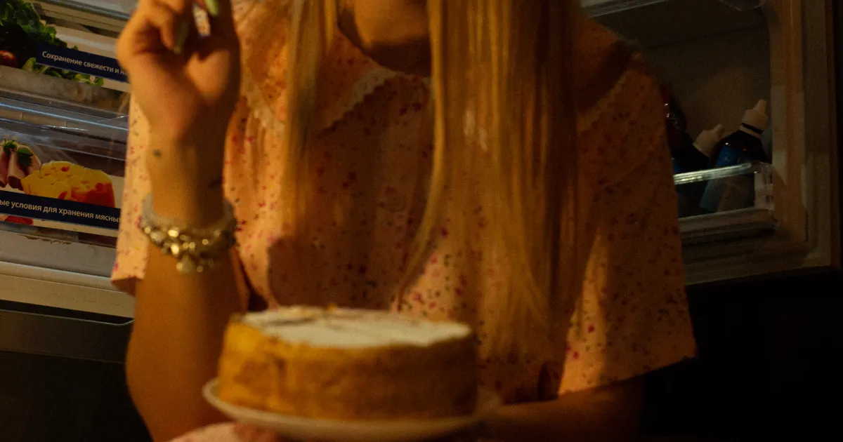 Someone eating a slice of cake in front of an open fridge at night