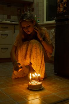 A woman crouching indoors lights candles on a birthday cake at night, creating a warm and intimate atmosphere.