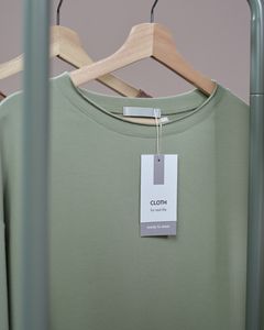 A simple green t-shirt with a tag hanging on a wooden hanger against a neutral background.