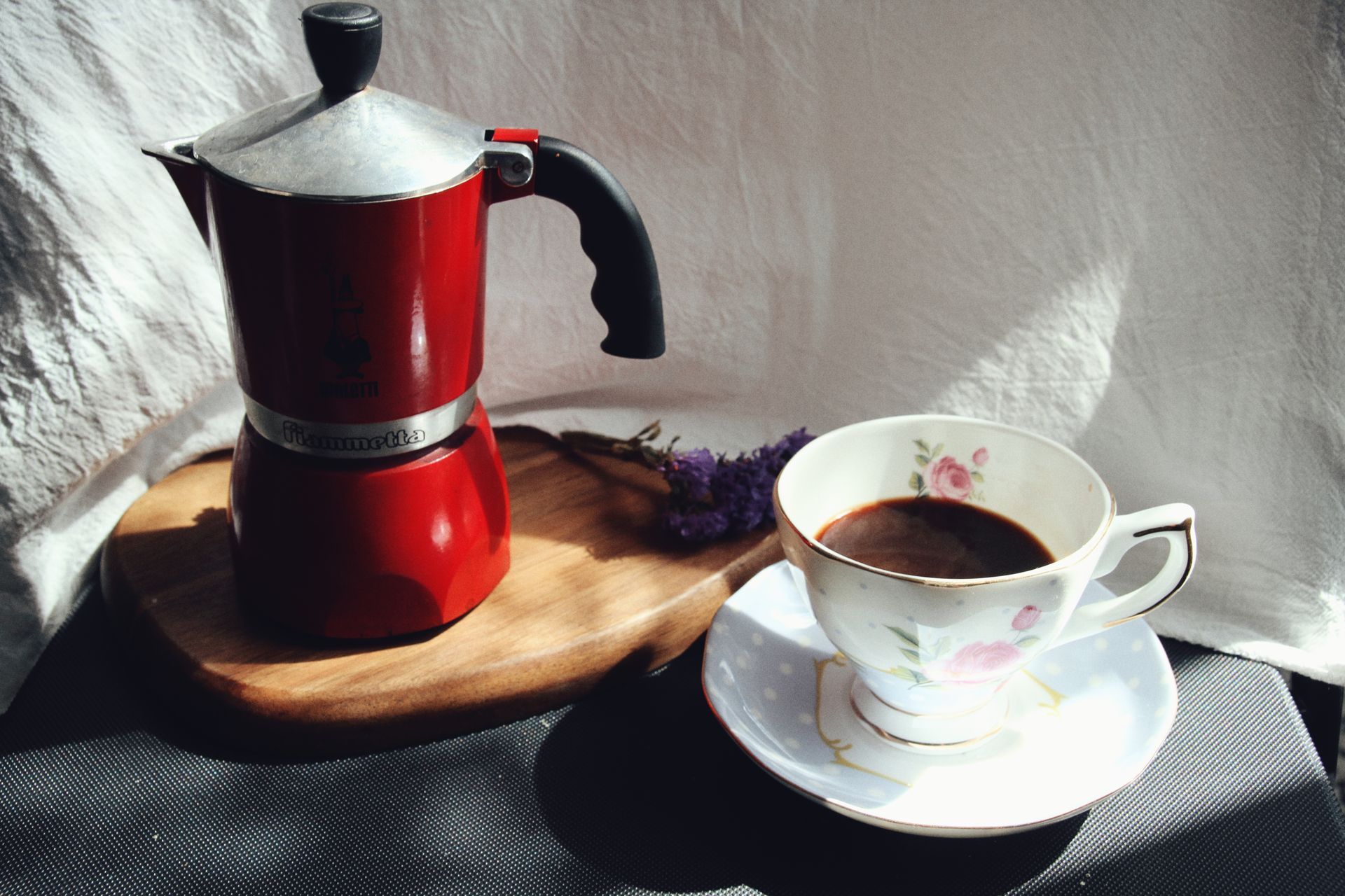 A cozy coffee scene featuring a red moka pot and floral teacup on a rustic board.