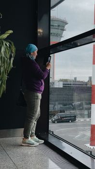 A woman stands inside as she takes photos through a large airport window on a cloudy day.