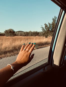 A hand with teal nail polish reaches out of a car window towards a scenic rural landscape.
