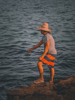 A man in a straw hat fishes from rocks by the sea at sunset, capturing coastal serenity.