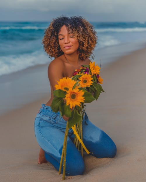 Woman in Denim Pants Kneeling on the Beach while Holding Flowers