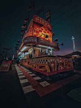 Illuminated pizza stand at night, serving drinks and cheeseburgers in an urban fair setting.