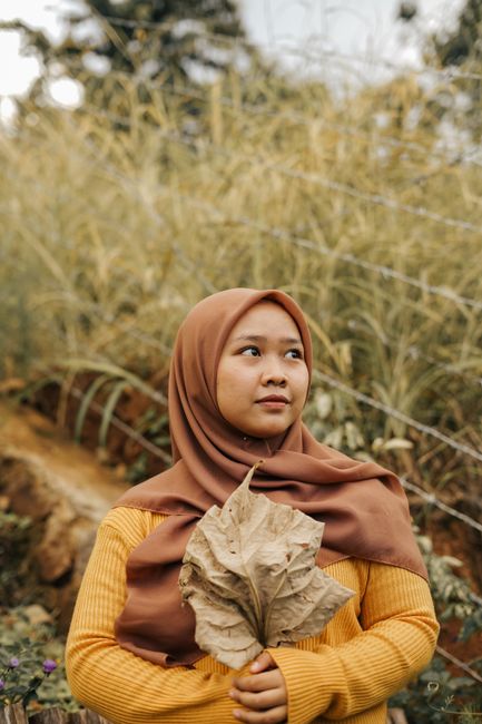 A young woman in a brown hijab holds a dried leaf in an outdoor setting, evoking a serene, earthy mood.