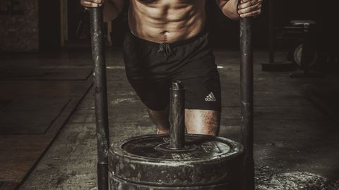 A shirtless man engages in a heavy sled push workout indoors, showcasing strength and endurance.