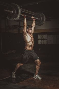 Shirtless man lifting barbell in a dimly lit gym, showcasing strength and fitness.