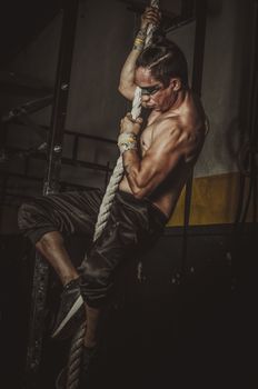 Strong man showcasing fitness and strength while climbing a rope indoors.