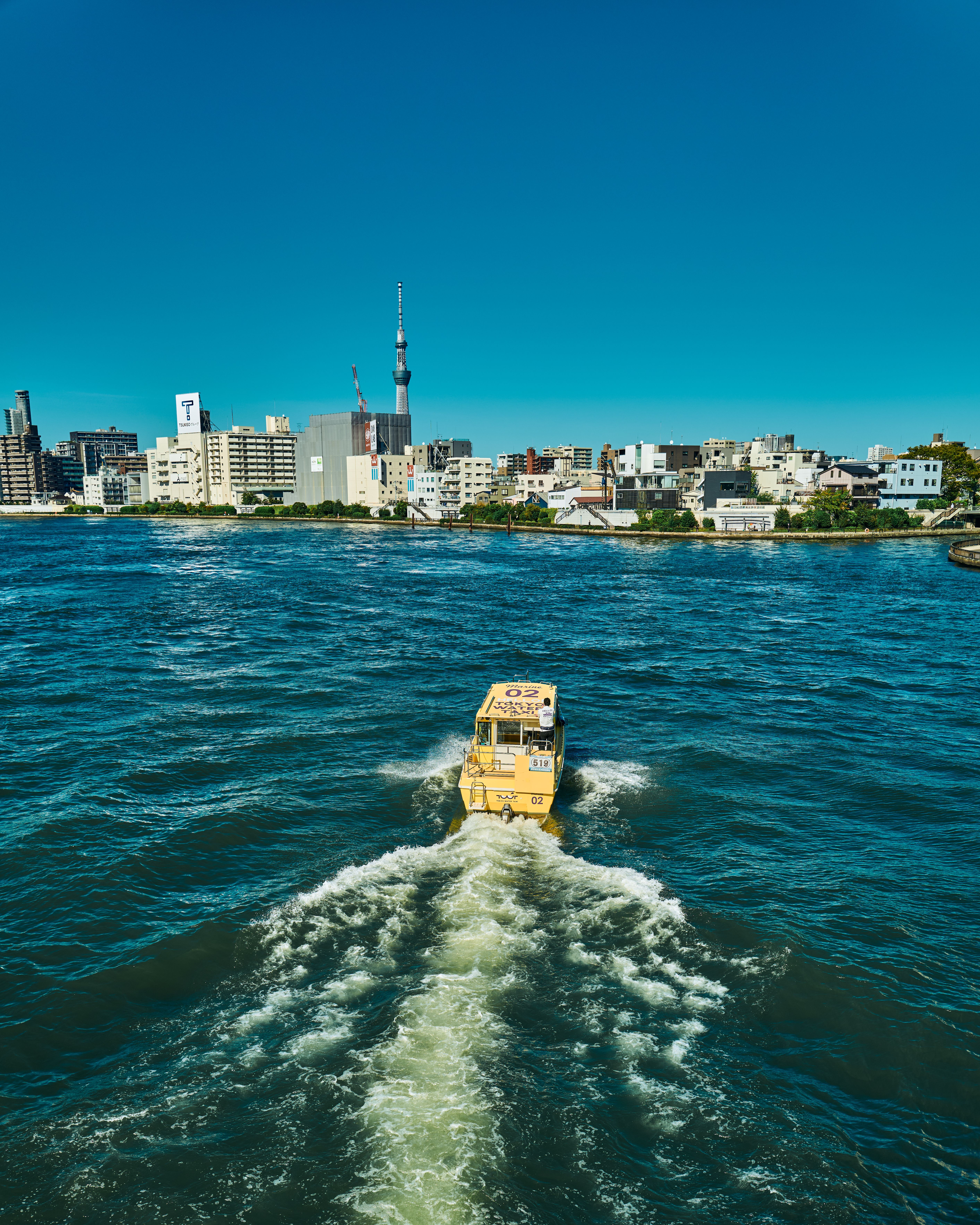 A vibrant city skyline with a yellow boat crossing a vivid blue river on a sunny day.