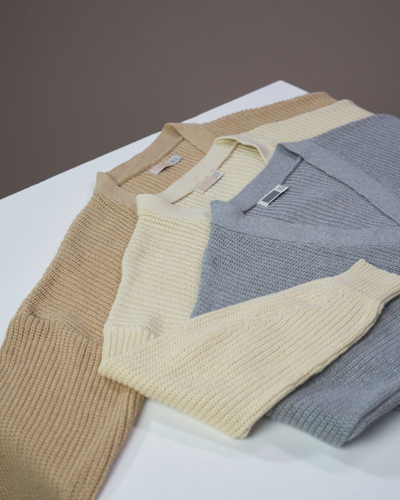 Tiga cardigan rajut warna netral ditata rapi di atas latar abu-abu