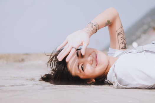 Asian woman with tattoos lying on the ground, smiling and covering her face from sunlight.