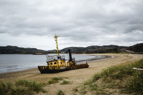Free A rusted shipwreck rests on a deserted beach under cloudy skies, creating a moody coastal scene. Stock Photo