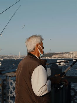 A man in a face mask fishes from a bridge with a city skyline in the background.