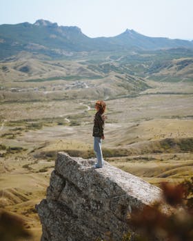 A woman stands on a rocky cliff overlooking a vast mountain landscape, embracing nature and adventure.