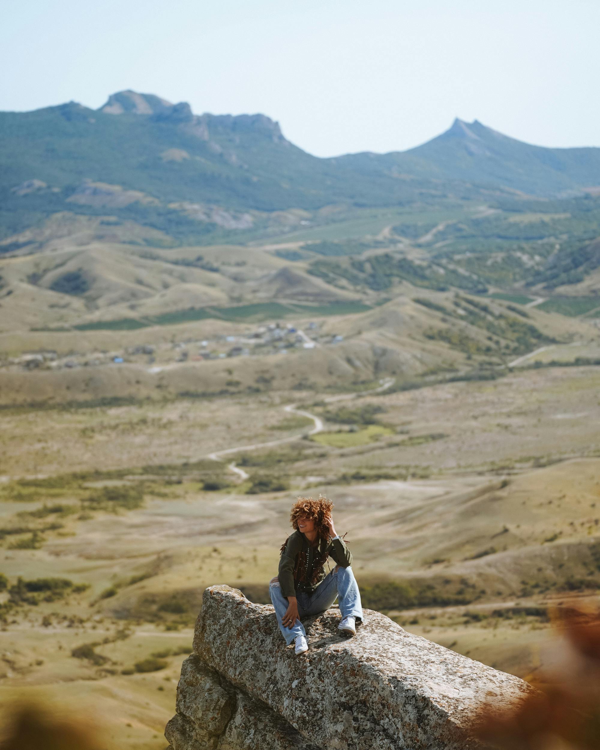Woman with curly hair sitting on rock, enjoying view of vast mountain landscape under clear sky.