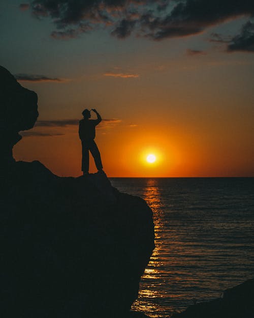 Silhouette of Man Standing on Rock Formation Near Body of Water during Sunset