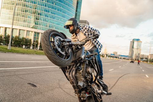 Free Biker in plaid shirt performing wheelie stunt on city street with modern buildings in background. Stock Photo