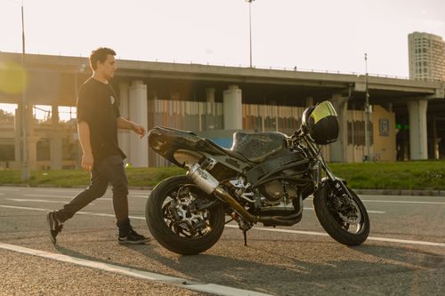 Free A man walks beside his motorcycle near a bridge during sunset in a city setting. Stock Photo