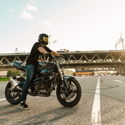Free A motorcyclist wearing a helmet rides on an urban road with a bridge in the background during sunset. Stock Photo