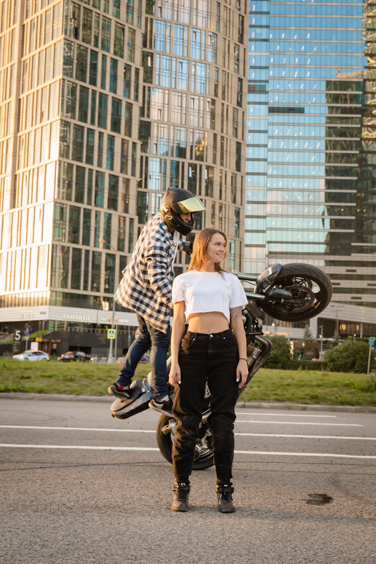 Woman In Crop Top Standing Beside A Man Riding A Motorcycle Doing A Stunt