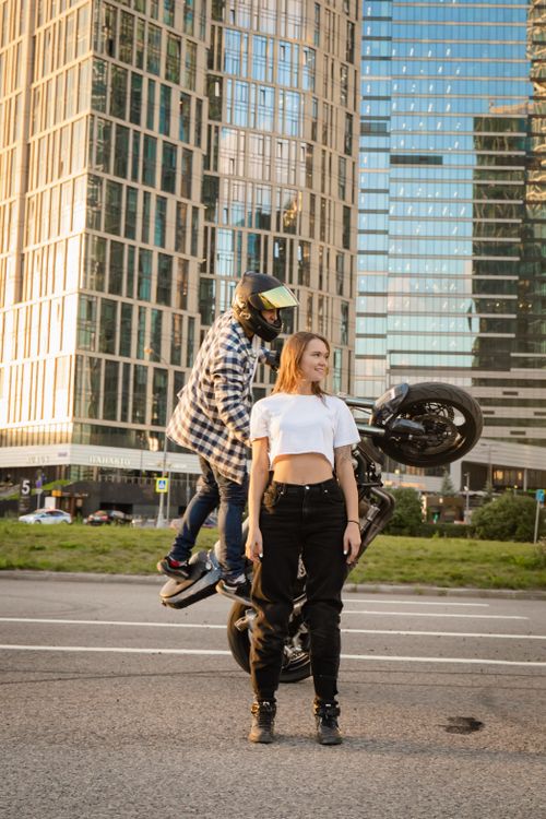 Free A motorcyclist performing a stunt on an urban road, framed by tall skyscrapers, with a woman standing nearby. Stock Photo