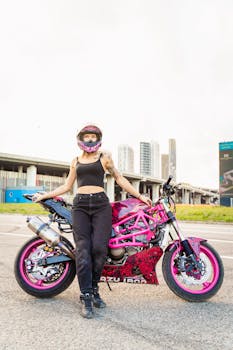 Confident woman standing with a pink custom motorcycle in an urban setting, wearing a helmet and casual attire.