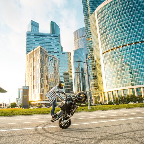 Free Motorcyclist does a wheelie in front of modern skyscrapers, showcasing urban thrill. Stock Photo