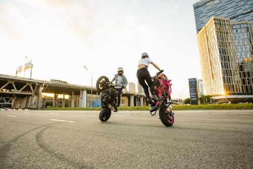 Free Two motorcyclists performing wheelies on city streets against a sunset backdrop. Stock Photo